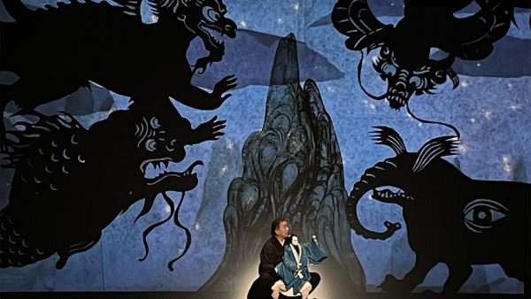 Photo of a man operating a puppet dressed in Japanese traditional clothes. The background is a stage set with a stylized mountain, a dark blue night sky, and menacing-looking fantastic beasts presented as shadow puppets.