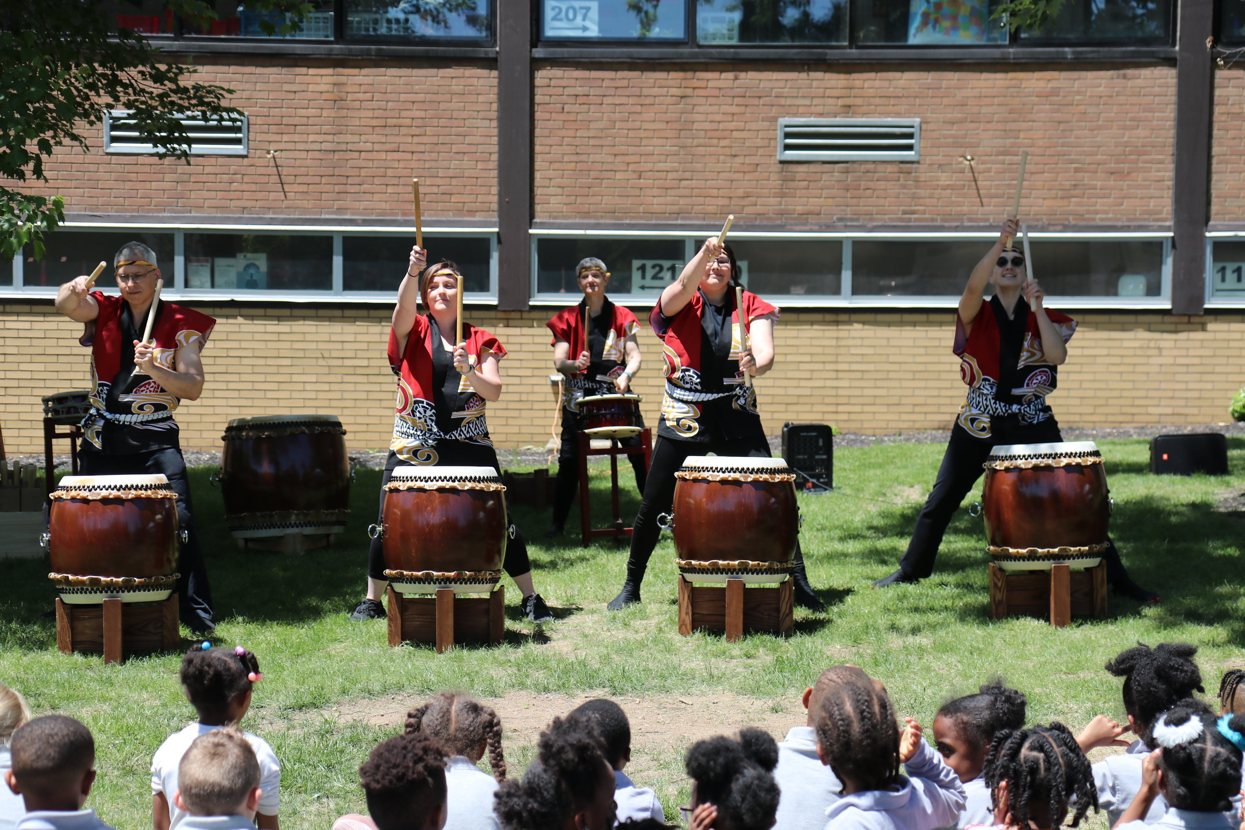 Pittsburgh Taiko members perform on a grass lawn in front of a school building. Several rows of elementary school students in uniforms are seen from behind, seated on the lawn facing the performers. Photo by Faruk Ozel.