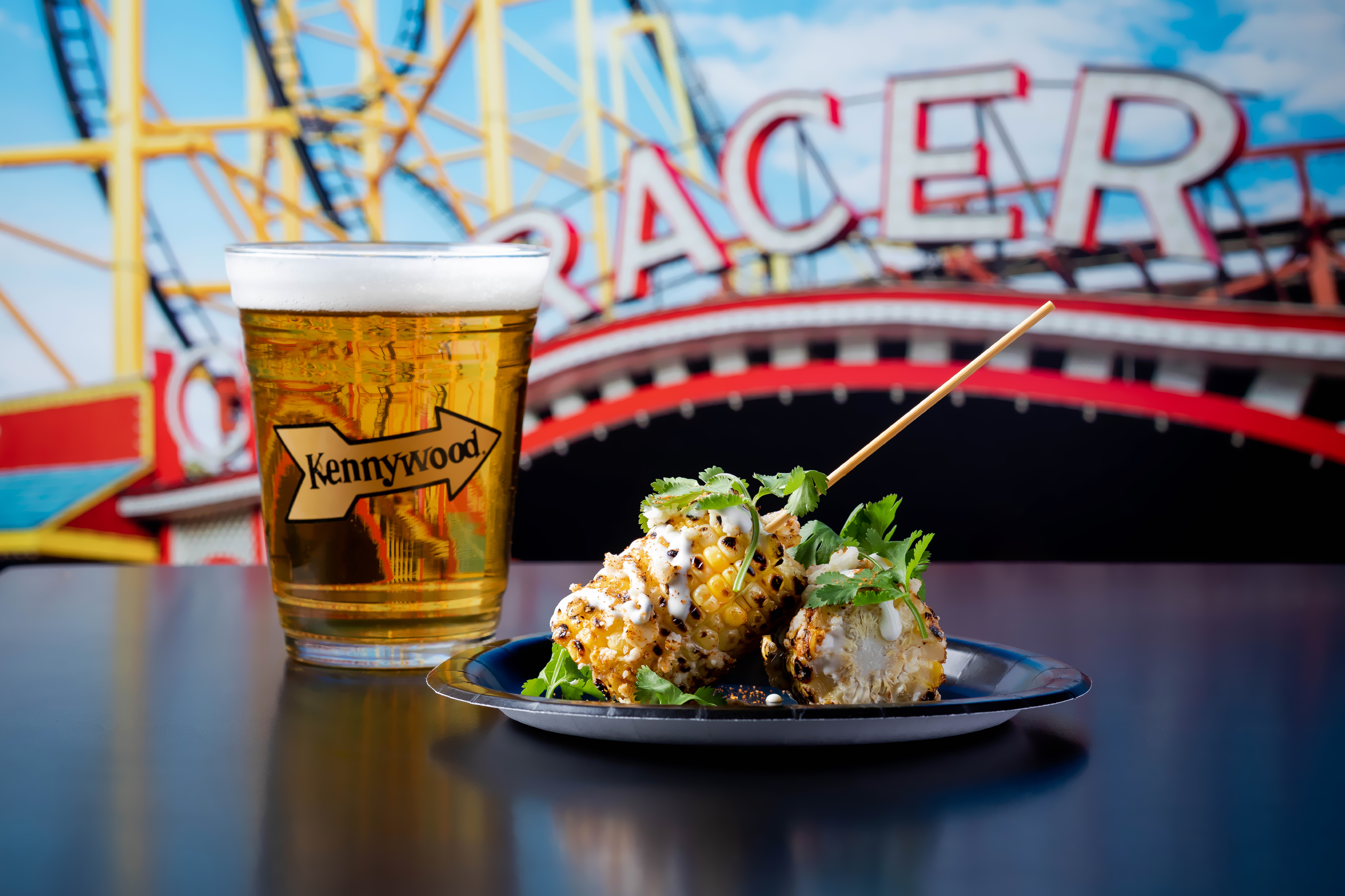 Photo of a draft beer in a clear plastic cup with a Kennywood logo, next to a plate of food with a fried coating, herb garnish, and a skewer, sitting on a dark table. In the background are roller coaster tracks and the sign for The Racer. 