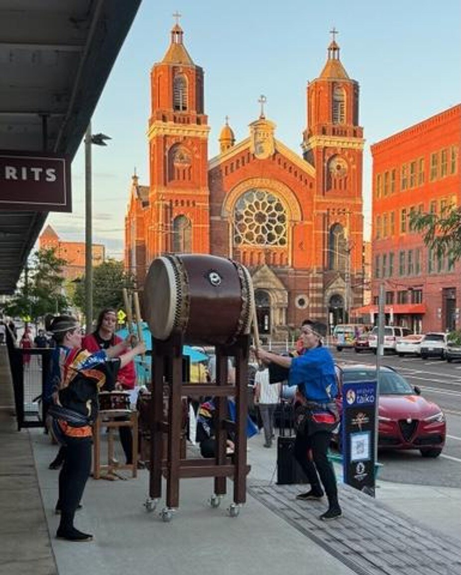 Photo of Pittsburgh Taiko playing odaiko on the sidewalk in front of Novo Food Hall in June 2024. Smallman Street is visible on the right side of the photo, and St. Stanislaus Kostka Church is centered in the background.