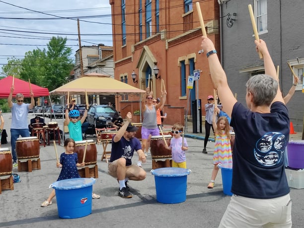 Photo of a Pittsburgh Taiko member leading a group of children and adults in a taiko drumming exercise. The drums are set up near a church in the middle of a closed street.