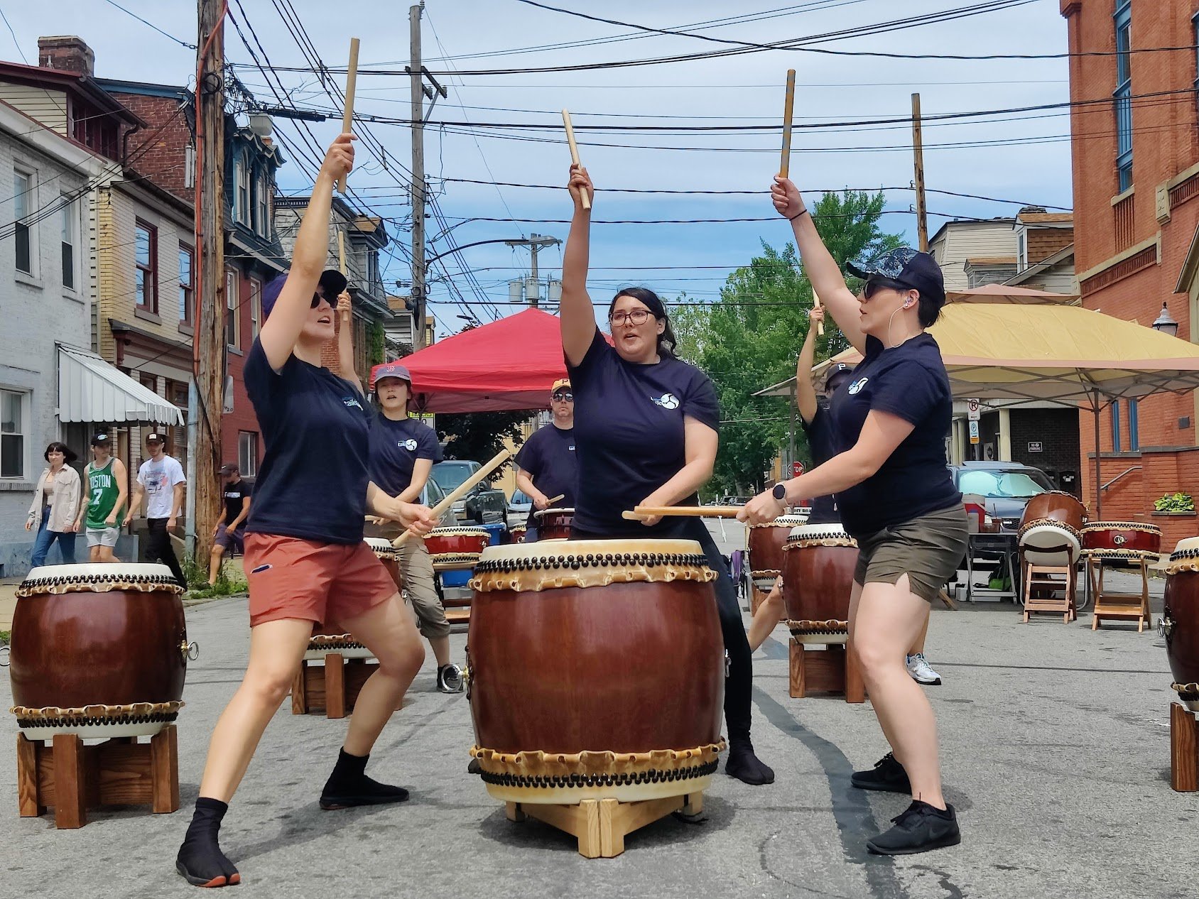 Photo of three Pittsburgh Taiko performers playing on a single drum, under a blue sky on a closed street in the South Side.