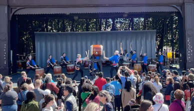Pittsburgh Taiko performing on an outdoor stage in front of a large crowd of people. It's a sunny day.