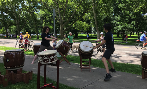 Two Pittsburgh Taiko members perform in a park while cyclists and dog walkers pass by on the road. From Open Streets 2017.