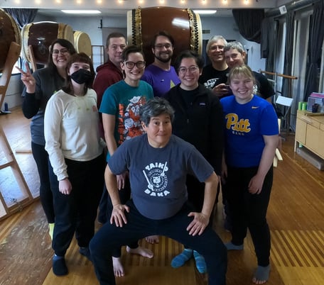 group photo of Pittsburgh Taiko members and Tiffany Tamaribuchi posing in front of a drum at the workshop.