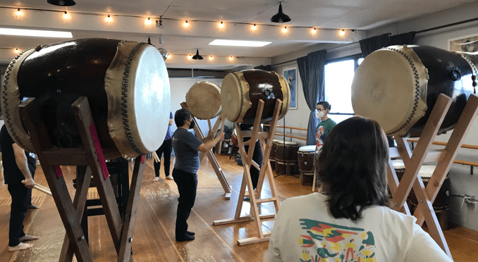 Casual photo of the workshop, showing several large taiko drums on upright stands, and students standing at the drums. Tiffany Tamaribuchi is at a drum explaining and demonstrating a point.